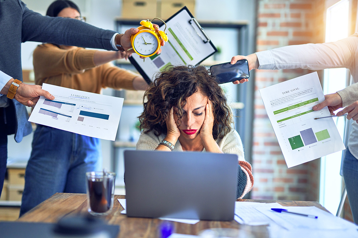 Woman at laptop looking stressed with people handing her papers