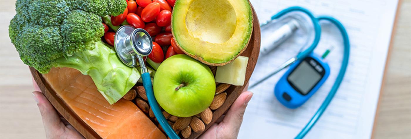 Person holding a heart-shaped bowl with fruits and vegetables. A stethoscope and blood sugar monitor are pictured as well.